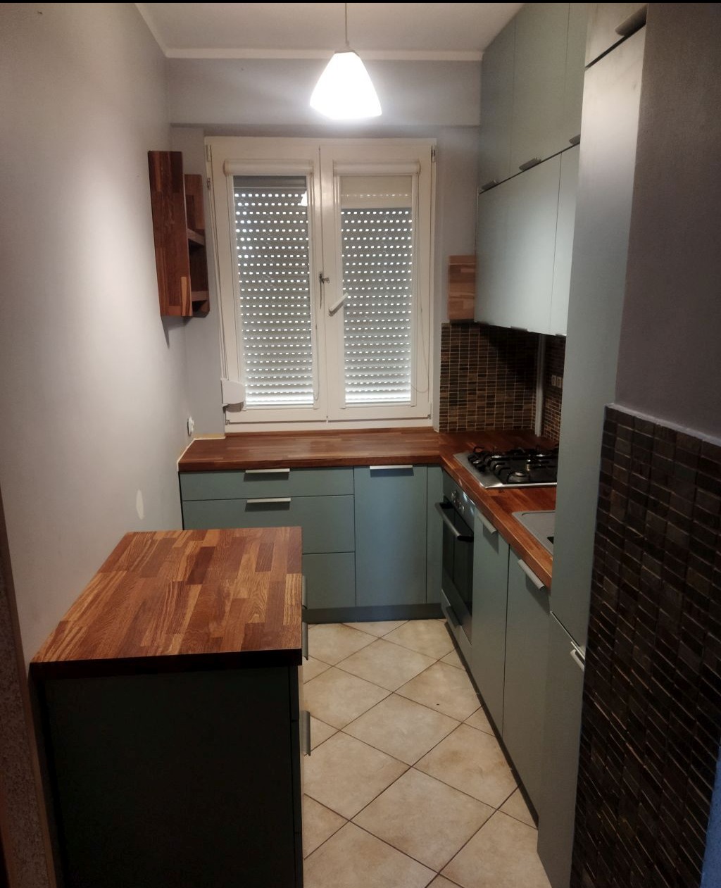 Niewielka kuchnia w bloku z zabudową w odcieniach szarości i drewnianymi blatami, widok na okno z roletami i brązowe płytki nad blatem.