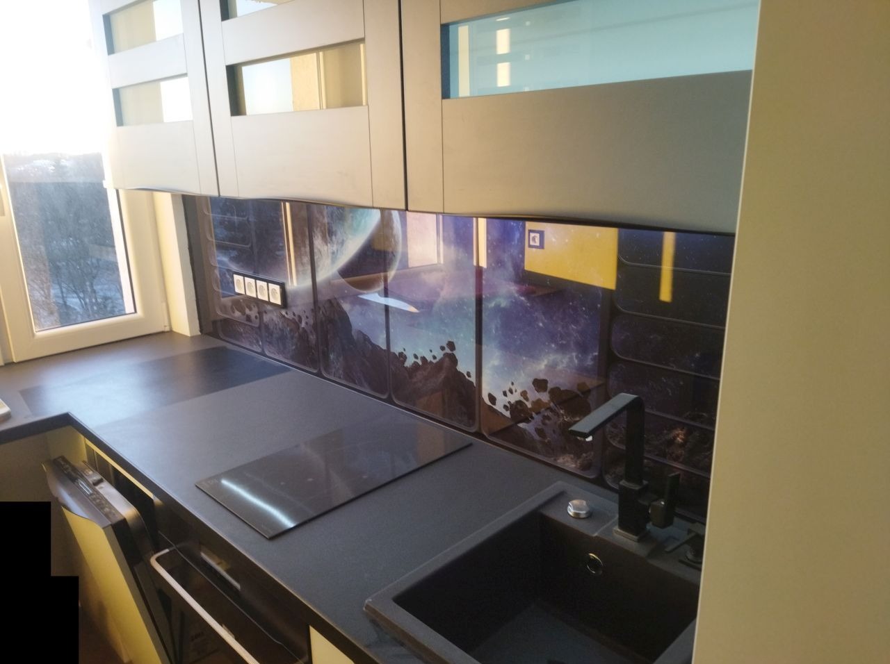 Nowoczesna kuchnia z czarnym blatem, płytą indukcyjną i zlewozmywakiem, wyróżniająca się panelem ściennym z motywem kosmicznym i widokiem na planetę.