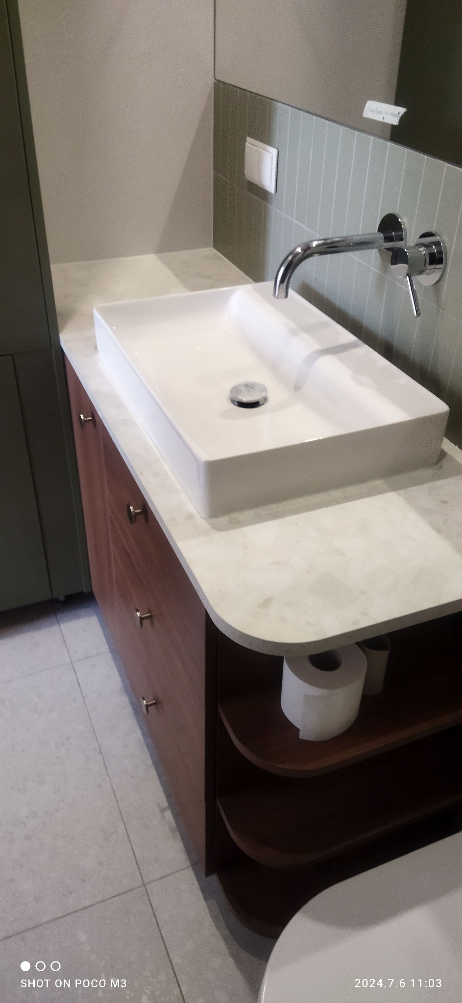 Nowoczesna biała umywalka nablatowa o kwadratowym kształcie, zamontowana na drewnianej szafce z szufladami i półkami, z widocznym fragmentem toalety w dolnym rogu.