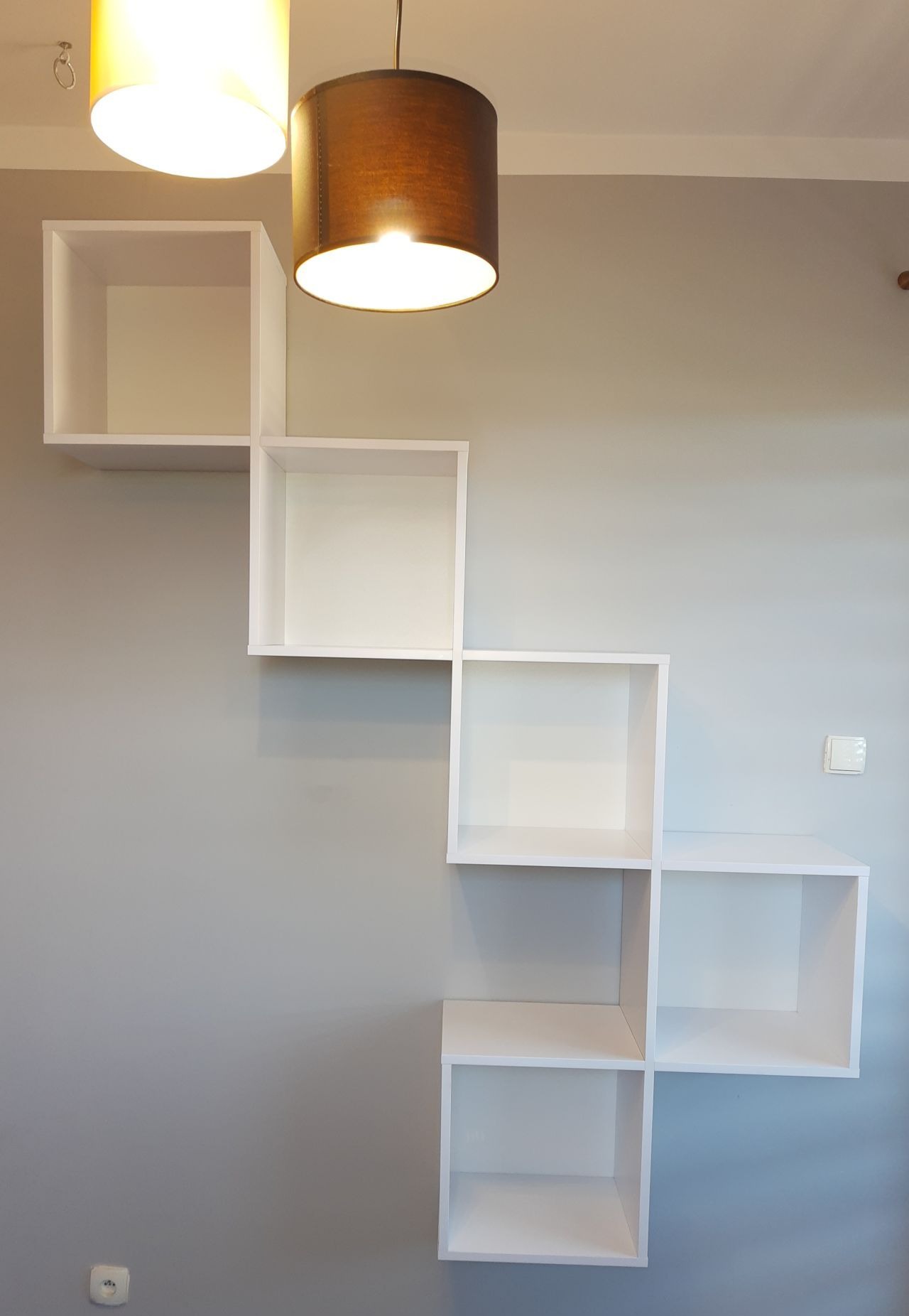 Białe, modułowe półki ścienne w kształcie schodów, oświetlone przez dwie lampy wiszące, na szarej ścianie z gniazdkiem i włącznikiem.