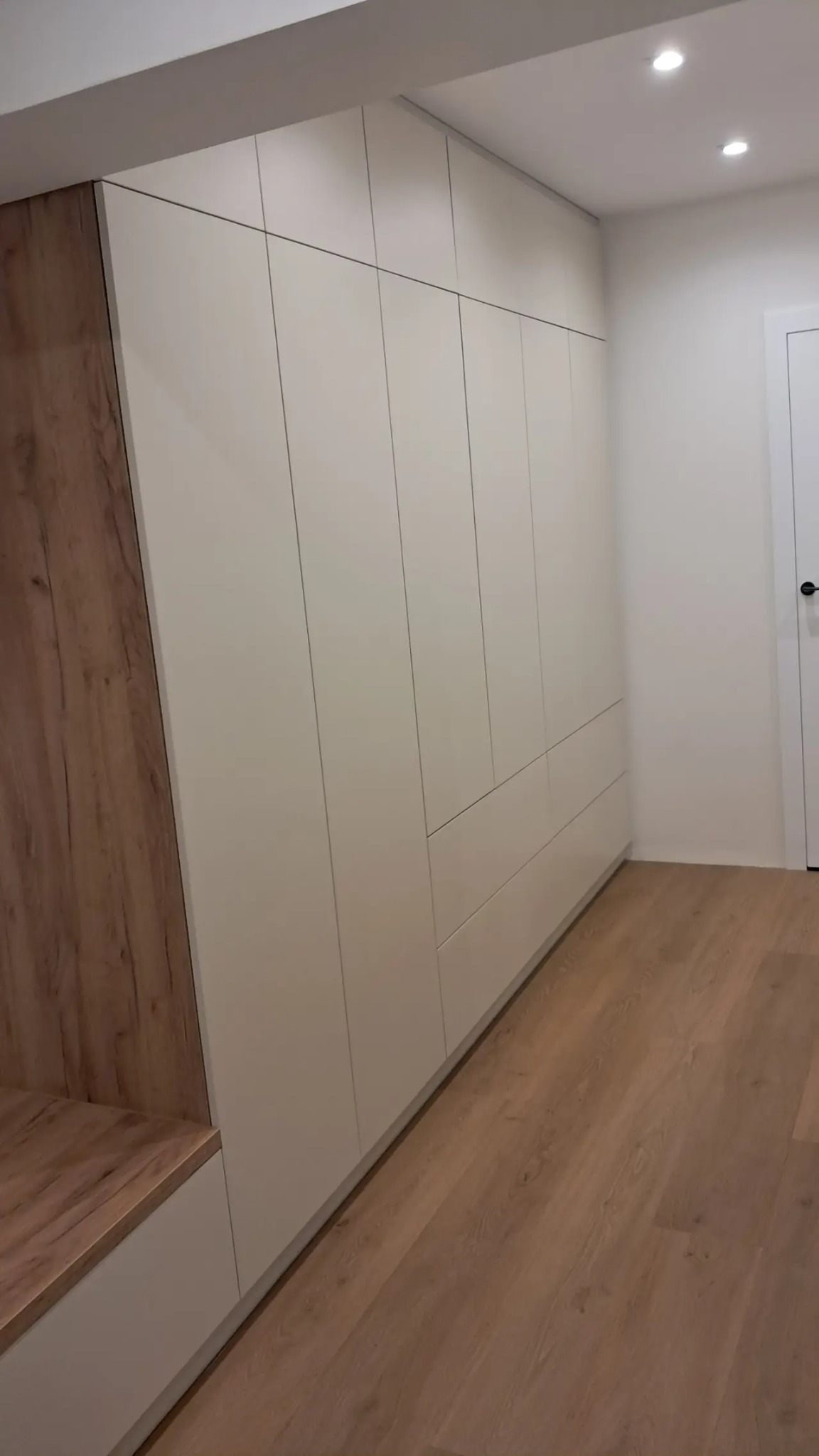 Biała szafa wnękowa z drewnianym akcentem i siedziskiem, dopasowana do drewnianej podłogi. Minimalistyczny design, nowoczesne wykończenie, widok z perspektywy podłogi.