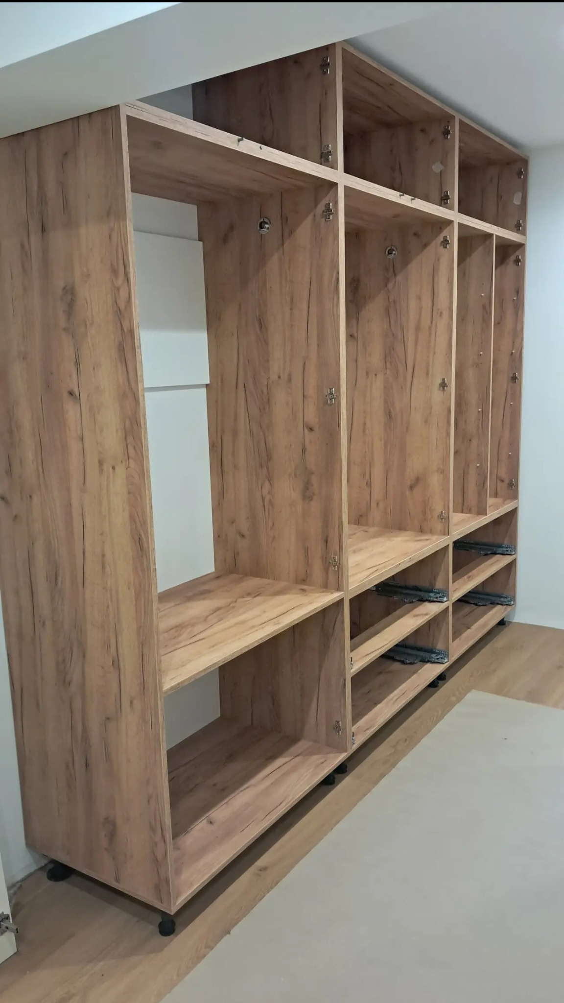 Szafa wnękowa z płyty drewnopodobnej, widoczne prowadnice szuflad i zawiasy. Ustawiona na podłodze z paneli, z regulowanymi nóżkami. Częściowo zamontowana.