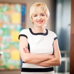 Portret uśmiechniętej blondynki w czarno-białej sukience, z założonymi rękami na tle abstrakcyjnego obrazu i przeszklonej ściany.