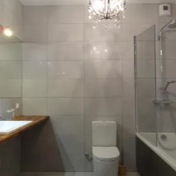 Nowoczesna łazienka z szarymi płytkami, drewnianym blatem pod umywalką i kryształowym żyrandolem, widok na toaletę i kabinę prysznicową za szklaną ścianką.