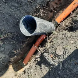 Rura drenarska karbowana czarna połączona z pomarańczową rurą kanalizacyjną w wykopie ziemnym, widoczne korzenie i grudki ziemi.