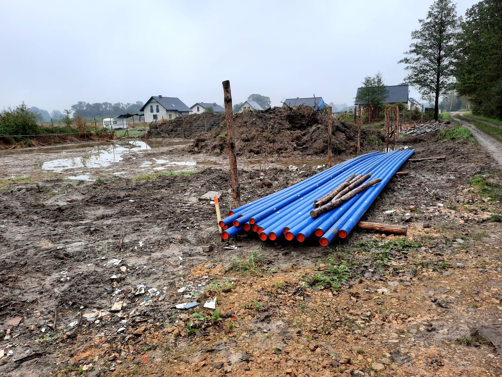 Stos niebieskich rur z pomarańczowymi końcówkami leżący na błotnistym gruncie obok wykopu, w tle domy i pochmurne niebo.