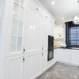 Biała, lakierowana zabudowa kuchenna z przeszklonymi witrynami i wbudowanym piekarnikiem, na podłodze szare płytki imitujące marmur.