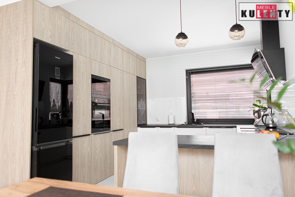 Nowoczesna zabudowa kuchenna z frontami w kolorze jasnego drewna, zintegrowana z czarną lodówką i piekarnikiem, oświetlona wiszącymi lampami z przydymionego szkła.