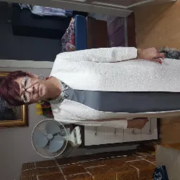 Kobieta w okularach i eleganckim, kremowym żakiecie pozuje w pomieszczeniu z terakotową podłogą, widoczny wentylator i obraz w złotej ramie.