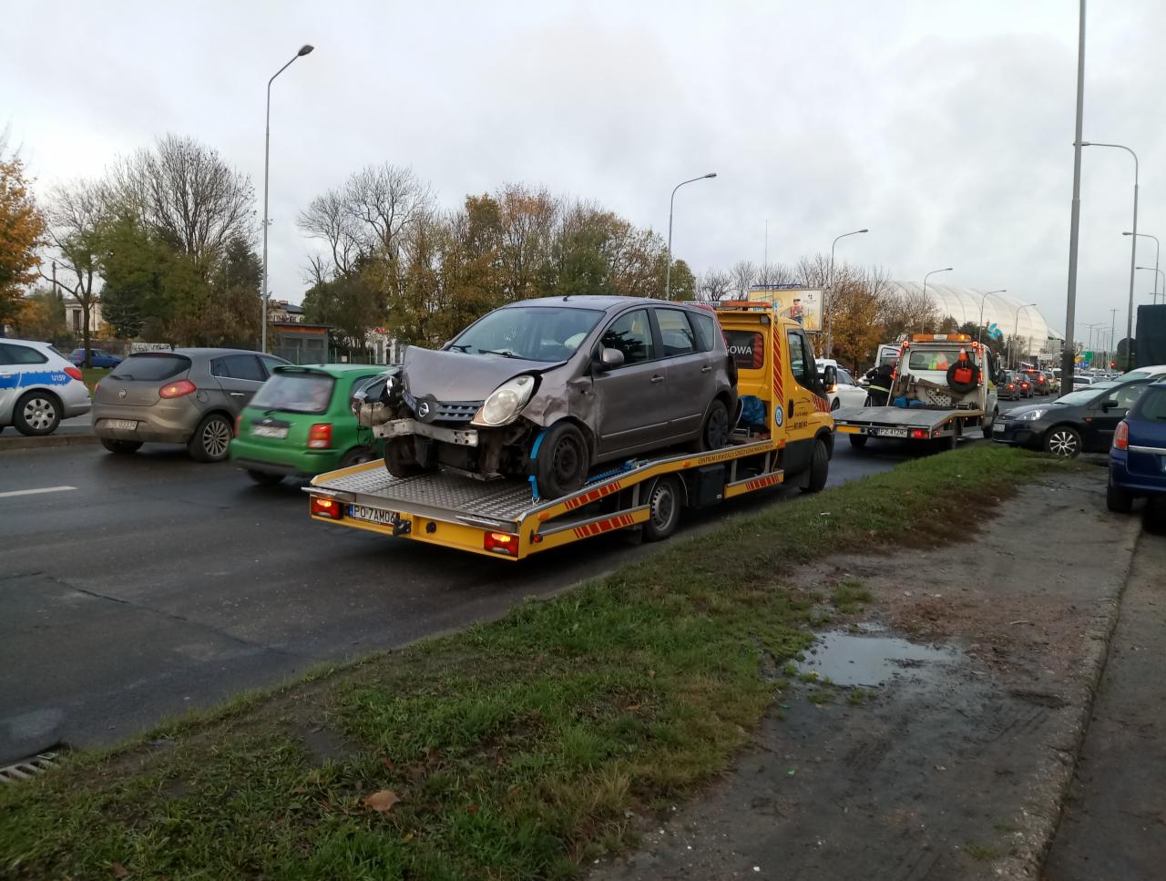 Uszkodzony, szary Nissan Note na żółtej lawecie w ruchu ulicznym, widoczne uszkodzenia przodu pojazdu, tablica rejestracyjna PO 7AM06.