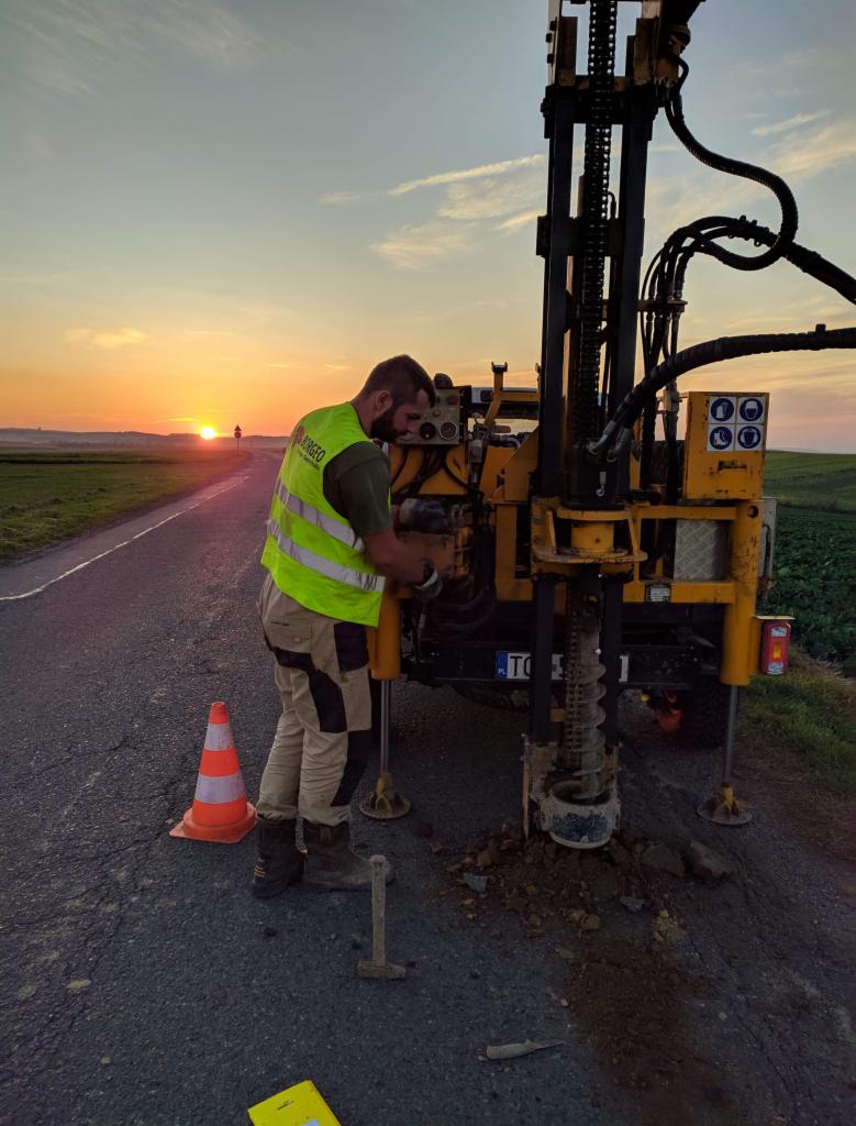 Pracownik w kamizelce odblaskowej obsługuje wiertnicę na poboczu drogi, w tle zachodzące słońce i krajobraz polny.