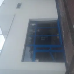 Biała elewacja budynku z dużym, niebieskim oknem; widoczna drabina na dachu i ceglana ściana z boku.