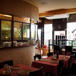 Wnętrze restauracji z ceglanym barem, szklanymi panelami z abstrakcyjnym wzorem, zastawionymi stołami z czerwoną kratą i widoczną kuchnią w tle.