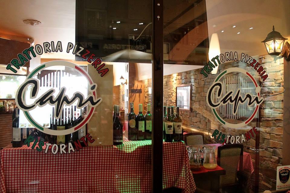 Wystawa przed wejściem do włoskiej restauracji z logo 'Capri', butelkami wina i dzbankiem lemoniady na czerwono-białym obrusie w kratkę, widok przez szklane drzwi.