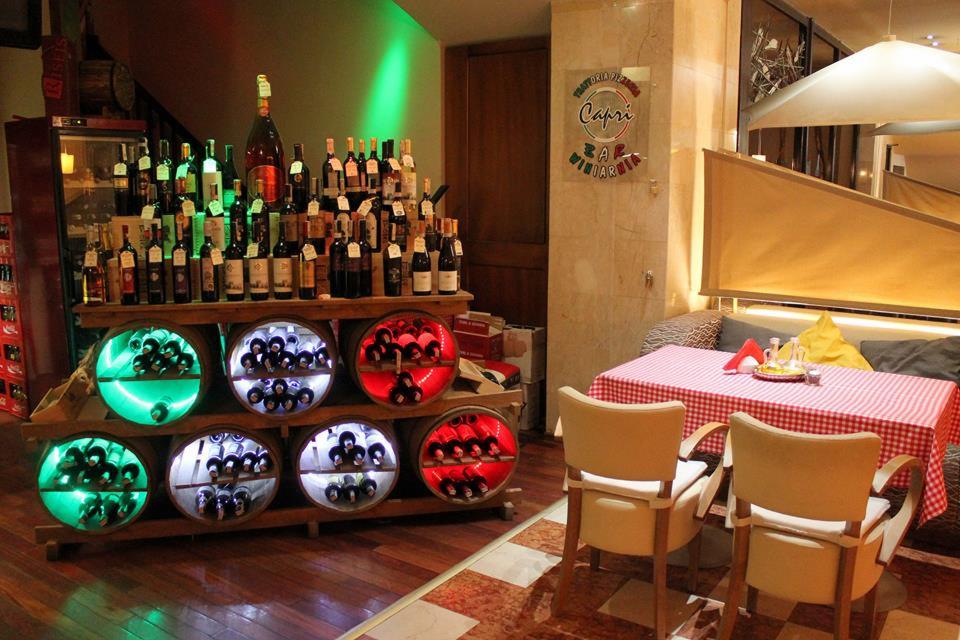 Wystrój restauracji: regał z podświetlonymi beczkami wina, butelki ułożone na górze, stolik z czerwoną kratą i kanapa w tle.