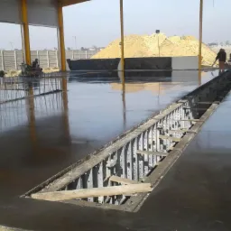 Świeżo wylana, gładka posadzka w hali przemysłowej, z widocznym kanałem rewizyjnym w trakcie budowy i stertą piasku na zewnątrz.