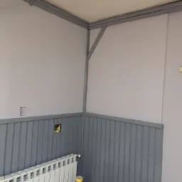 Świeżo pomalowane na szaro wnętrze z panelami ściennymi, widoczne puszki z farbą, grzejnik i niezamaskowane otwory w ścianie przed wykończeniem.