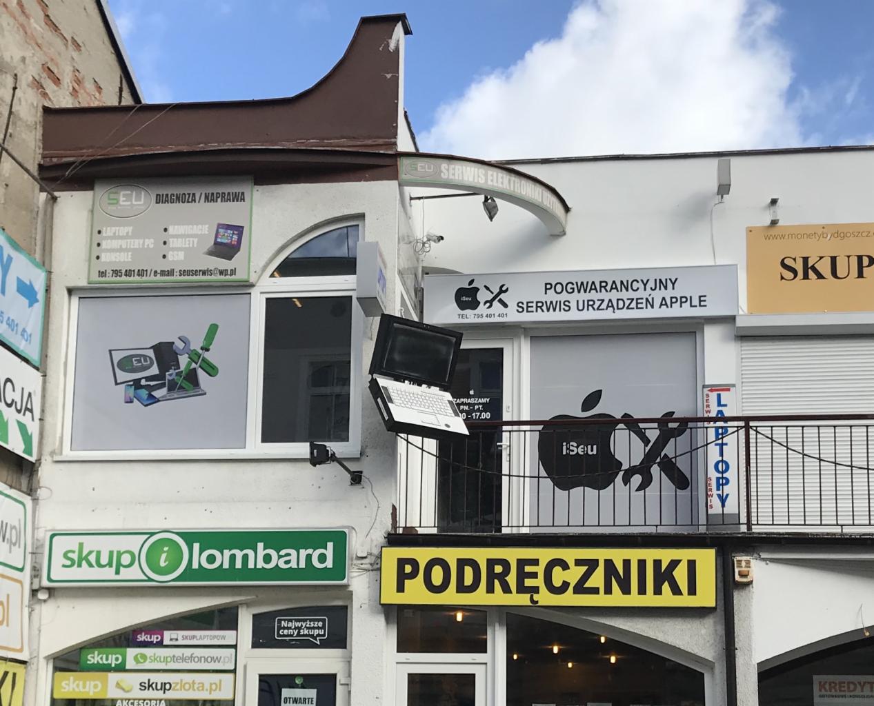 Fasada budynku z dwoma punktami serwisowymi: jeden oferuje diagnozę i naprawę laptopów, komputerów, tabletów i konsol, drugi pogwarancyjny serwis urządzeń Apple, z laptopem na zewnątrz zawieszonym...