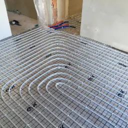 Białe rury ogrzewania podłogowego ułożone na siatce w surowym pomieszczeniu z widocznymi przewodami w tle, przygotowane do zalania wylewką.