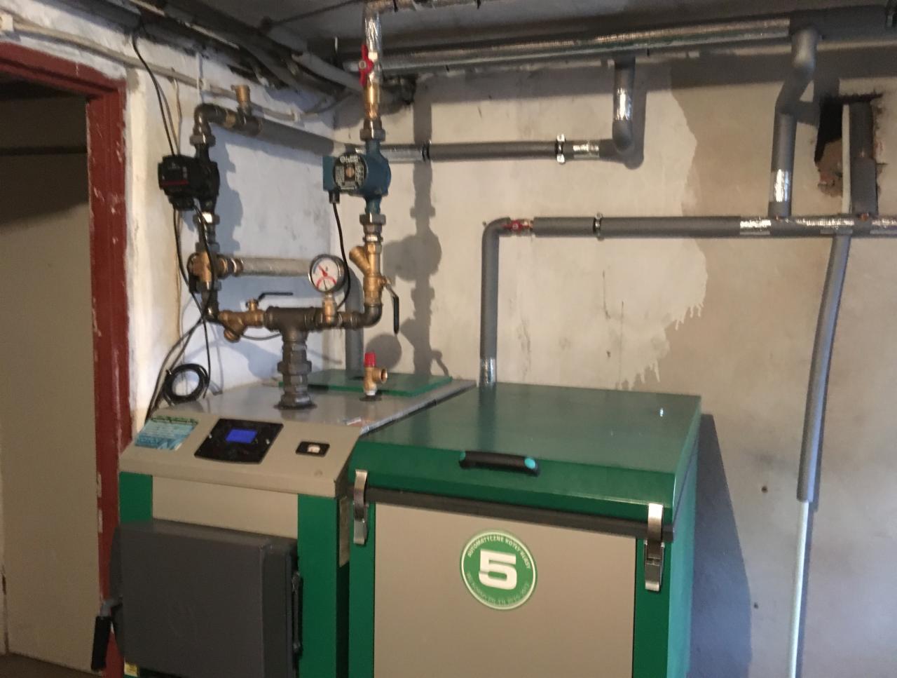 Nowoczesny piec na paliwo stałe z zielonymi akcentami, podłączony do instalacji hydraulicznej w piwnicy, widoczne rury, zawory i manometr.