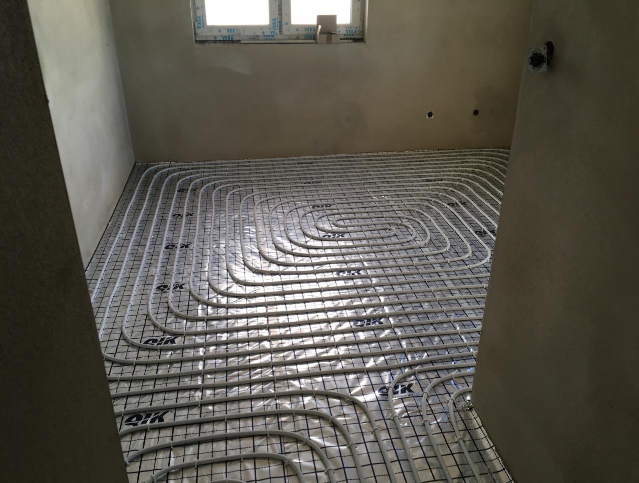 Białe rury ogrzewania podłogowego ułożone spiralnie na czarnej siatce montażowej, widoczne w surowym pomieszczeniu z oknem i szarą ścianą.