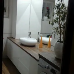 Nowoczesna łazienka z białą szafką pod umywalką z blatem drewnianym, okrągłą umywalką nablatową, lustrem, rośliną doniczkową i pralką.