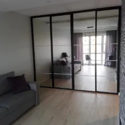 Nowoczesna szafa z lustrzanymi drzwiami przesuwnymi w czarnych ramach, odbijająca wnętrze salonu z szarą sofą i drewnianą podłogą.