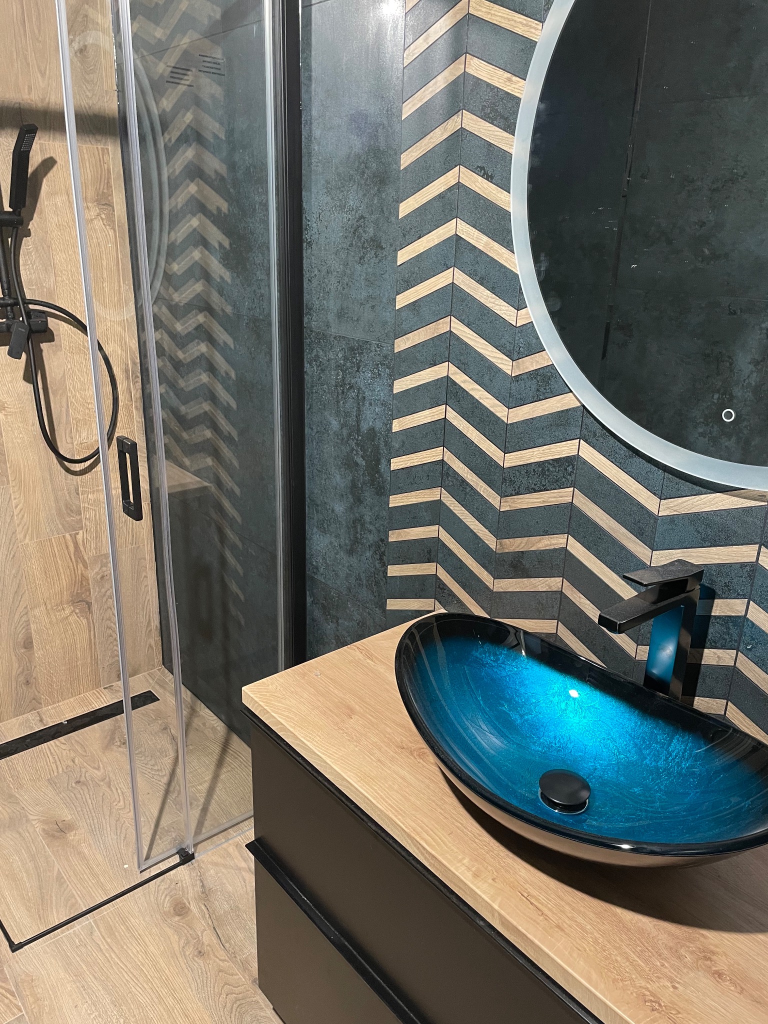 Nowoczesna łazienka z kabiną prysznicową typu walk-in, umywalką nablatową w odcieniach turkusu i baterią w kolorze grafitu, na tle ściany z płytek ułożonych w jodełkę.