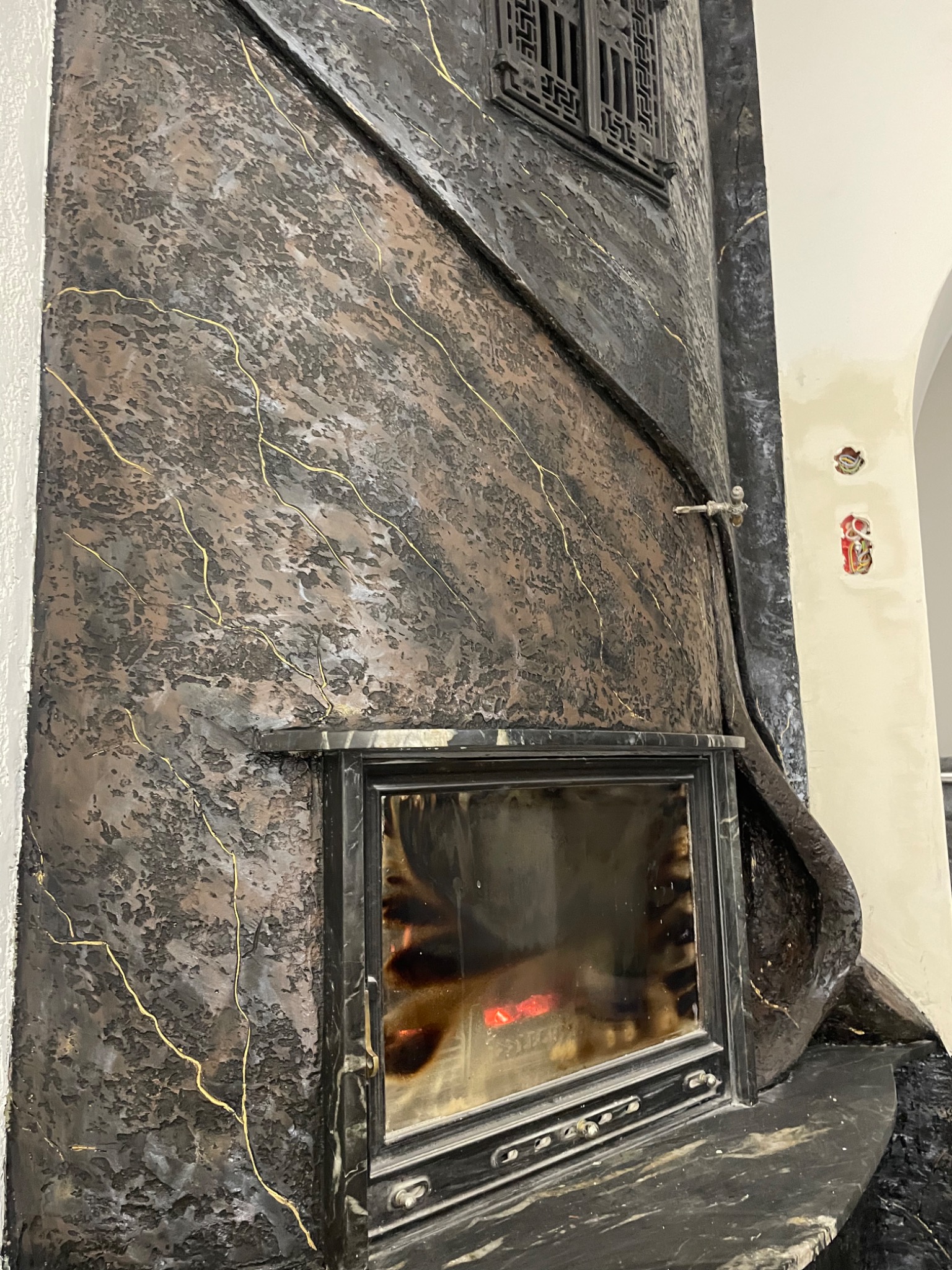 Ozdobny kominek z czarnego, teksturowanego tynku z efektem złotych żył, wkomponowany w ścianę z widocznymi elementami instalacji elektrycznej.