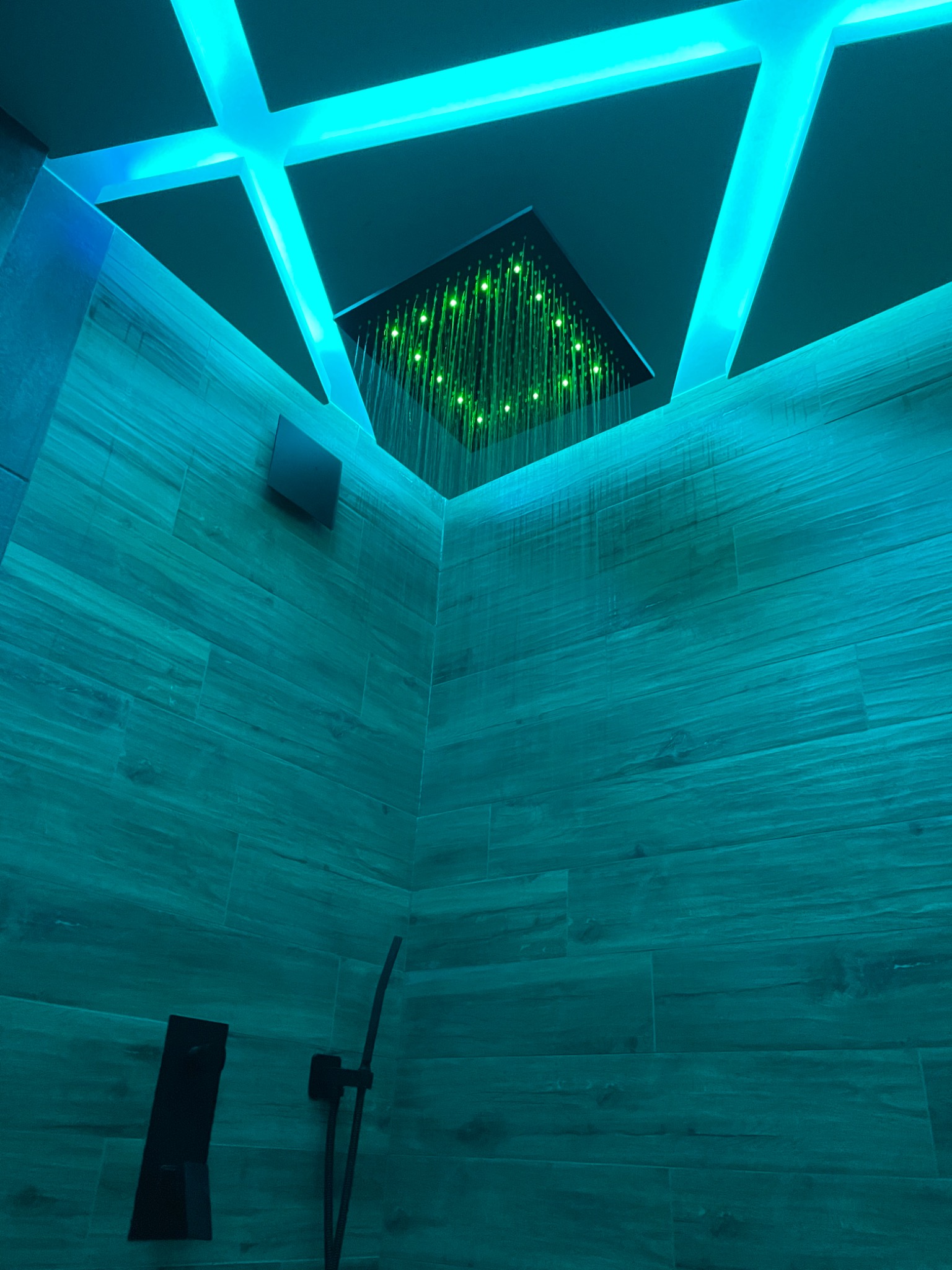 Nowoczesna kabina prysznicowa z deszczownicą LED, podświetlona turkusowym światłem, ściany wyłożone płytkami imitującymi drewno, czarna armatura.