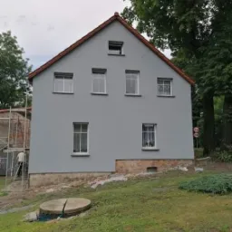 Dwukondygnacyjny dom z szarą elewacją i czerwonym dachem, z widocznymi śladami prac wokół fundamentów i rusztowaniem z boku.