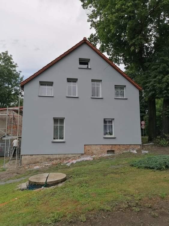 Dwukondygnacyjny dom z szarą elewacją i czerwonym dachem, z widocznymi śladami prac wokół fundamentów i rusztowaniem z boku.