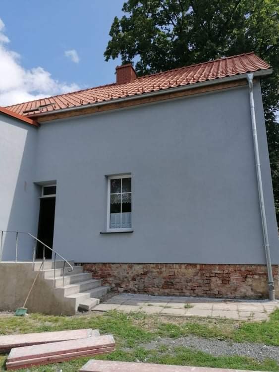 Świeżo otynkowana elewacja domu jednorodzinnego w odcieniach szarości, z widocznym fragmentem muru z czerwonej cegły przy podstawie i nową rynną, z drewnianym podbitką dachową i widocznymi schodami...