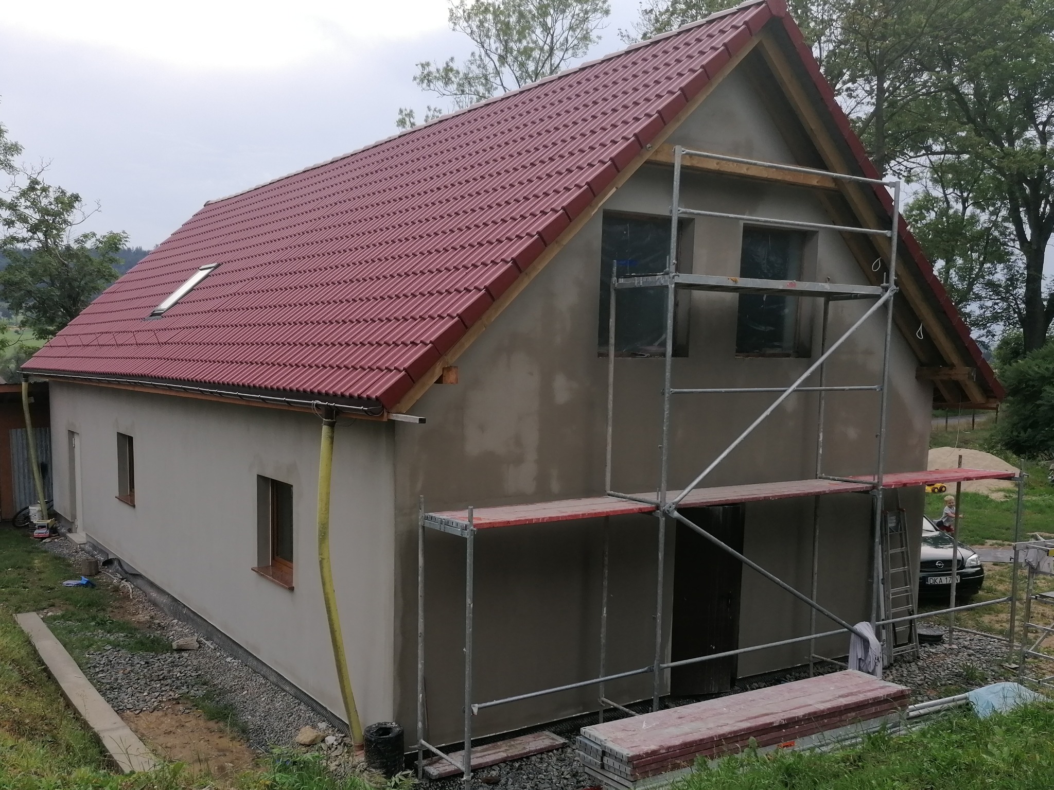 Dom jednorodzinny w trakcie prac elewacyjnych: surowa elewacja, czerwona dachówka, rusztowanie przy ścianie frontowej, widoczne okna zabezpieczone folią.