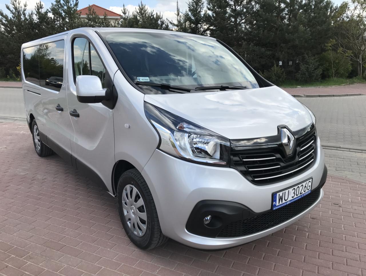 Srebrny Renault Trafic z polską tablicą rejestracyjną WU 3026G, zaparkowany na chodniku z cegły, z widocznym odbiciem chmur w przedniej szybie.