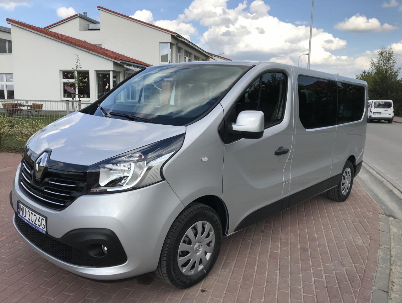 Srebrny bus Renault Trafic z przyciemnianymi szybami, zaparkowany na chodniku z kostki brukowej, na tle budynku z czerwoną dachówką i błękitnego nieba z chmurami.