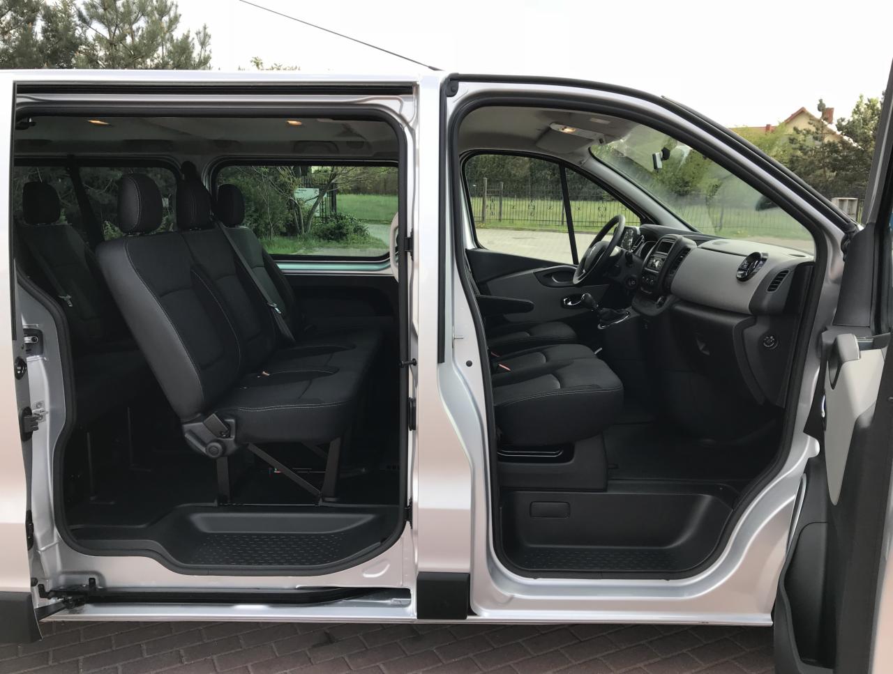 Wnętrze srebrnego busa z otwartymi drzwiami bocznymi, widoczne czarne fotele pasażerskie i kokpit kierowcy.