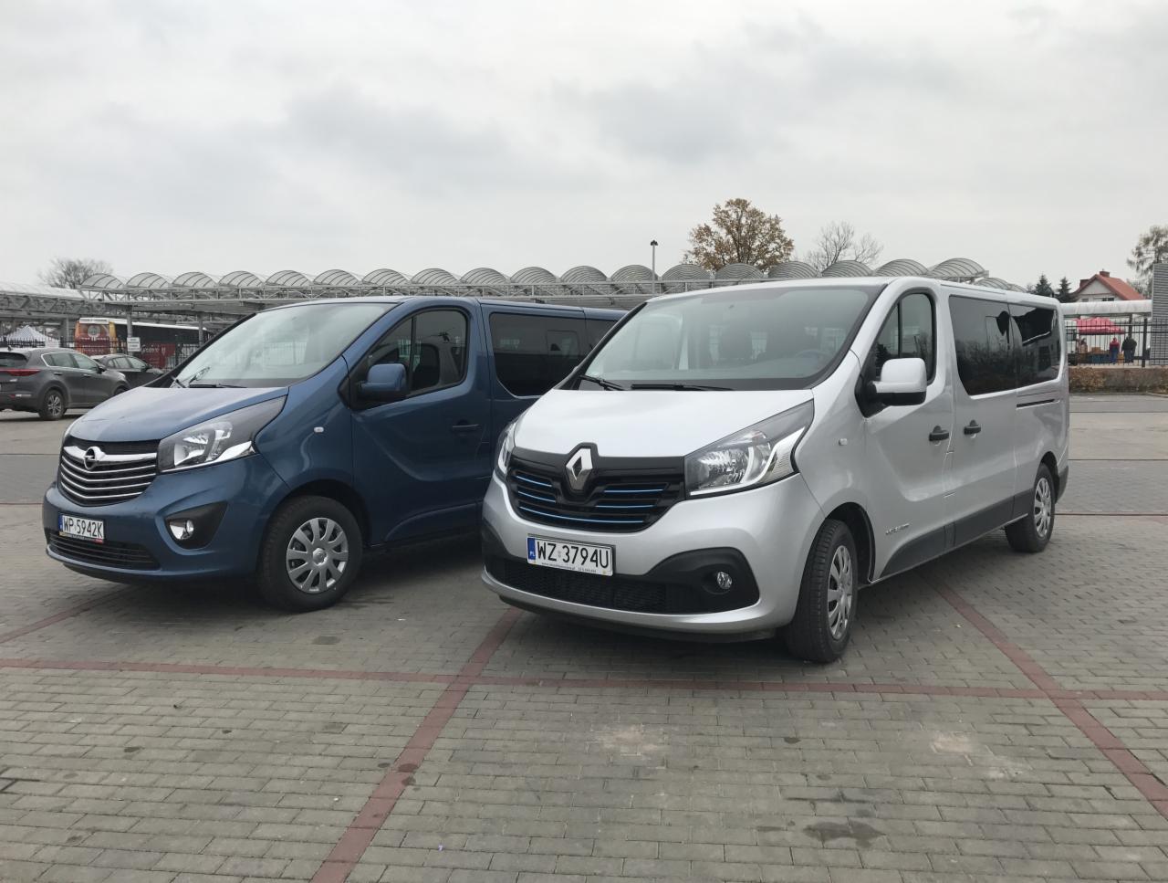 Dwa busy: niebieski Opel Vivaro i srebrny Renault Trafic, zaparkowane na brukowanym placu przed zadaszoną konstrukcją, pochmurne niebo w tle.