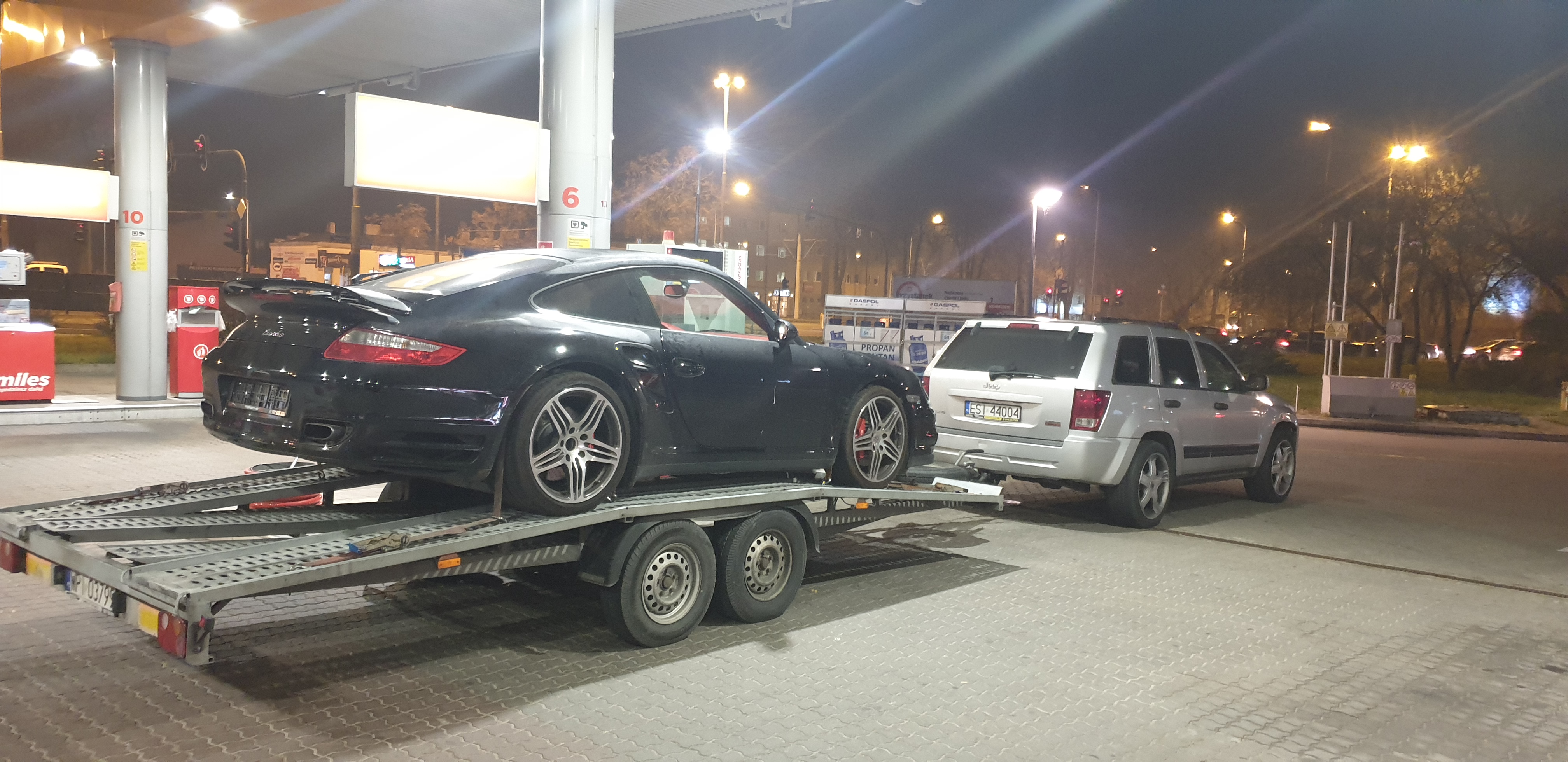 Czarny sportowy samochód Porsche na lawecie, holowany przez srebrnego Jeepa na stacji benzynowej nocą.