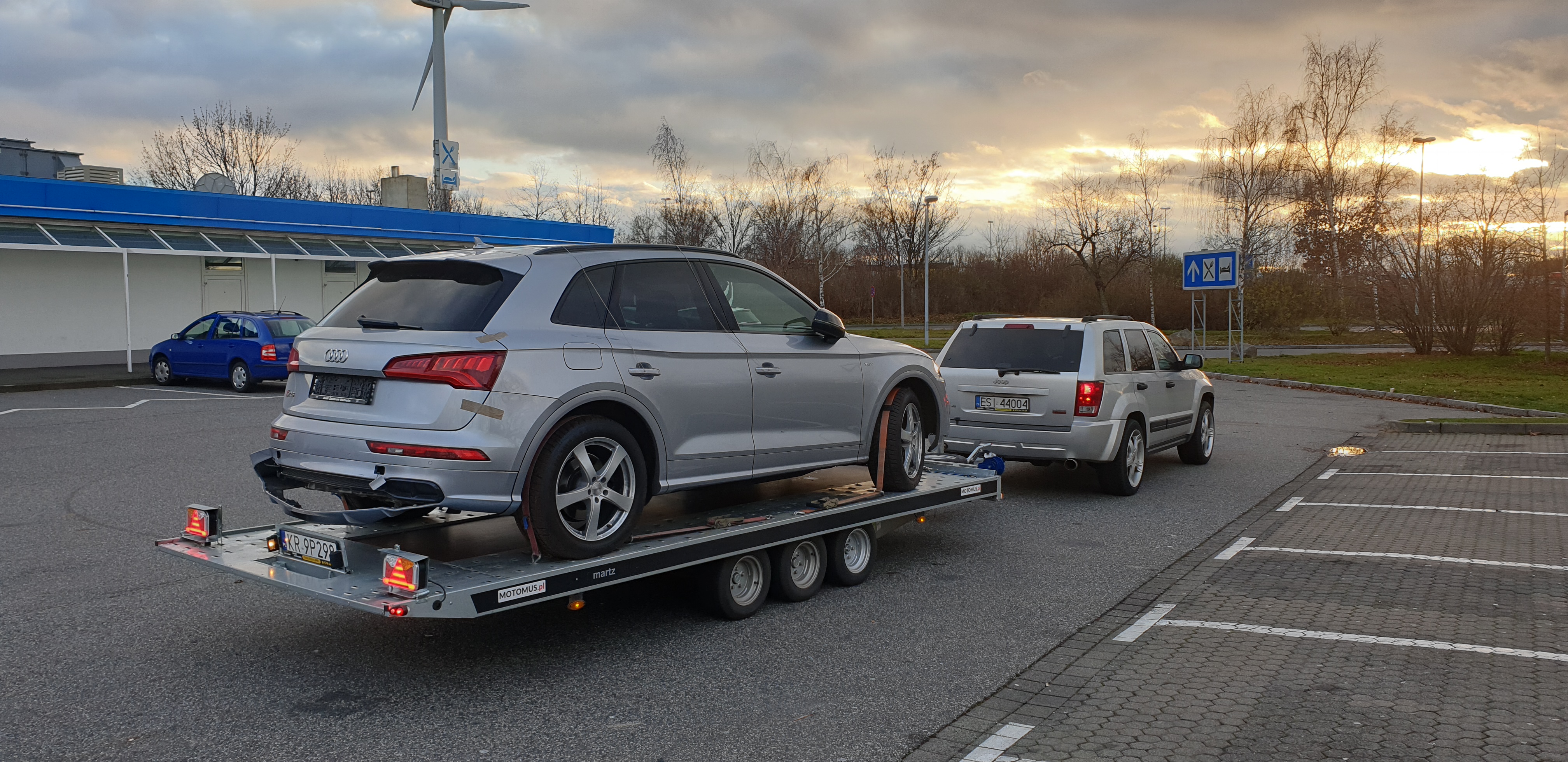 Srebrny uszkodzony Audi Q5 z taśmą na karoserii, zabezpieczony pasami na lawecie, holowany przez srebrnego Jeepa Grand Cherokee na parkingu z wiatrakiem w tle, rejestracje europejskie.
