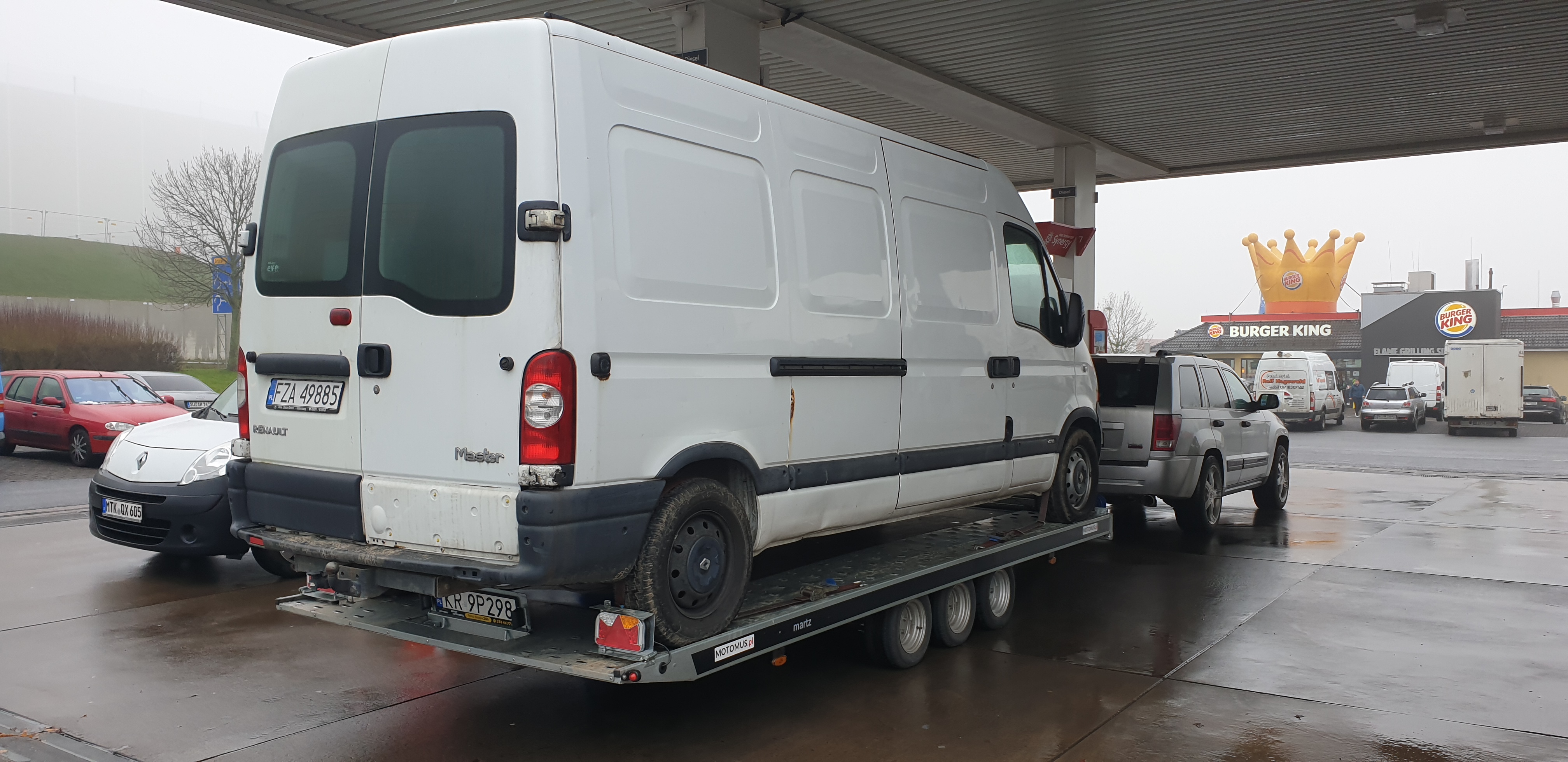 Biały bus Renault Master na lawecie typu platforma z trzema osiami, zaparkowany na stacji benzynowej z widocznym w tle Burger Kingiem z dużą koroną, widok z tyłu.