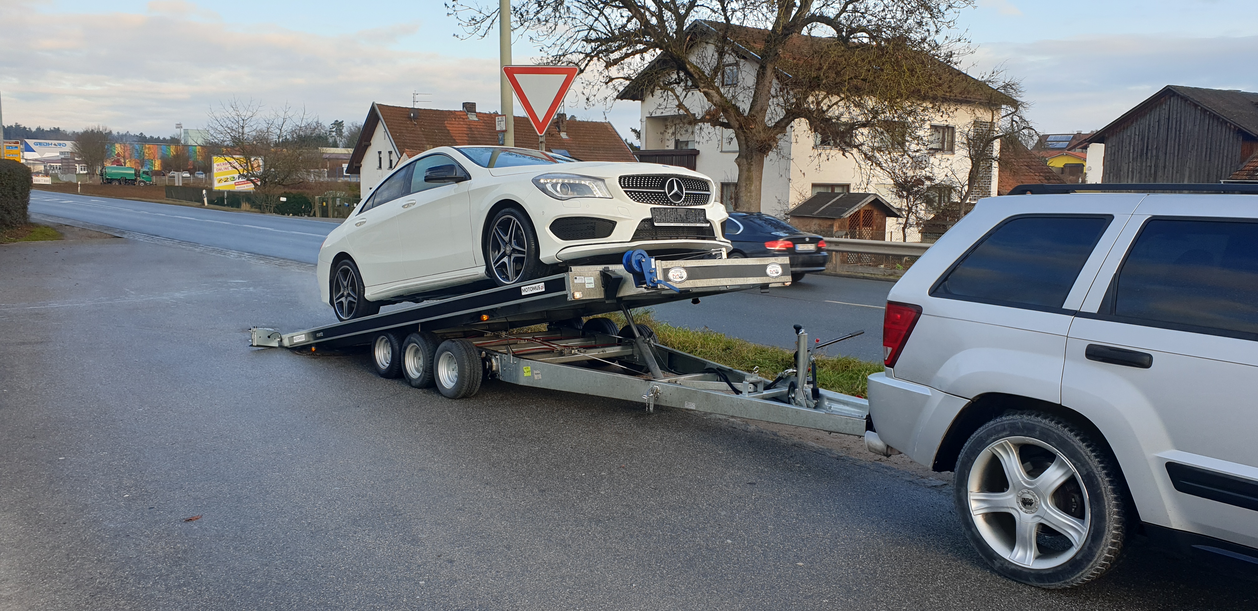 Biały Mercedes-Benz CLA na lawecie z napisem MOTOMUS.pl, holowany przez srebrnego Jeepa Cherokee na tle zabudowań wiejskich i znaku drogowego.