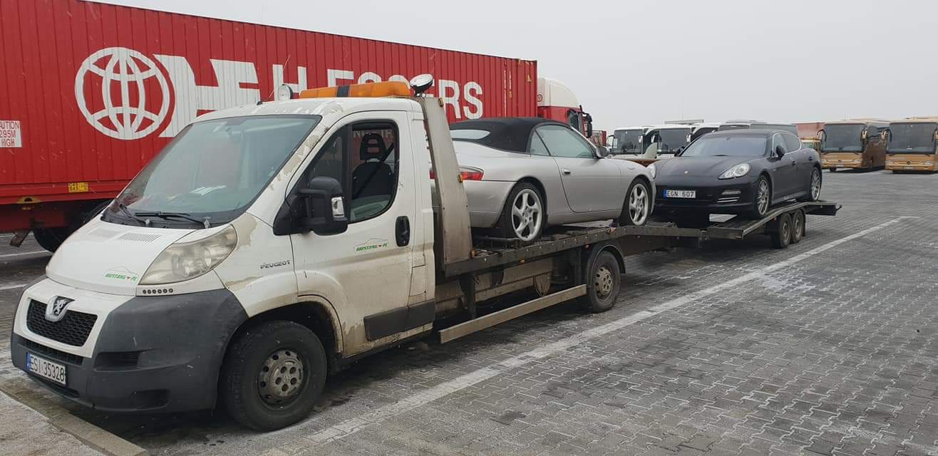 Biała laweta Peugeot z pomarańczowym kogutem transportuje srebrnego Porsche 911 Cabrio i czarne Porsche Panamera na dwuosiowej przyczepie w szary, pochmurny dzień, z widocznym czerwonym kontenerem...