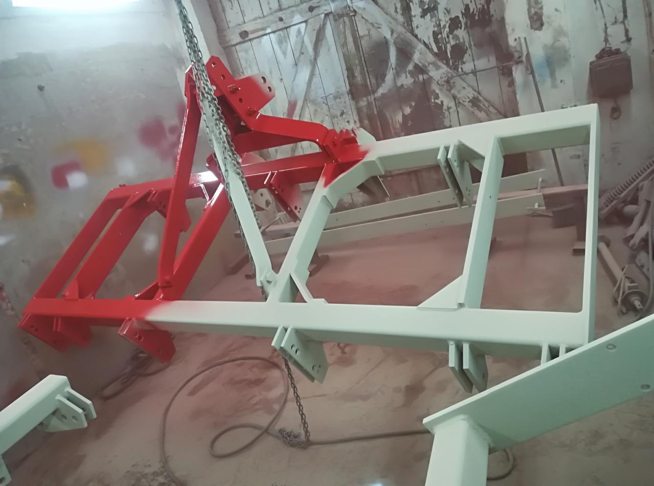 Metalowa konstrukcja rolnicza zawieszona na łańcuchach, częściowo pomalowana na czerwono, częściowo na biało, widoczna w warsztacie z narzędziami w tle.