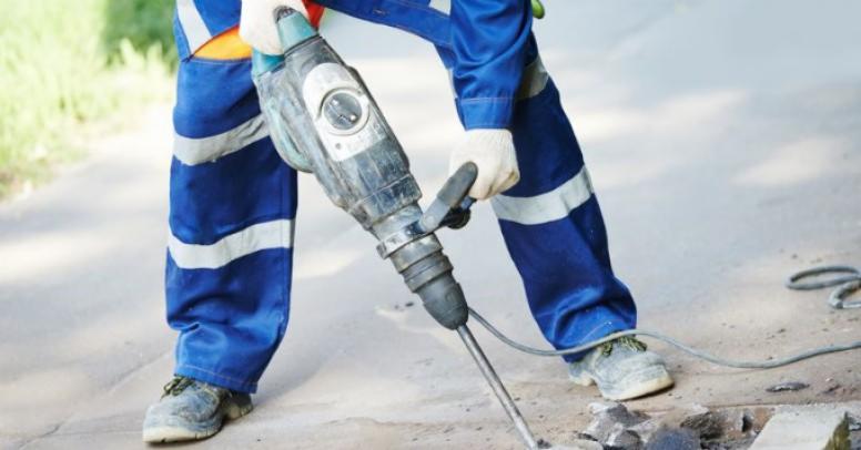 Pracownik w niebieskim kombinezonie z odblaskowymi pasami używa młota pneumatycznego do rozbijania betonowej nawierzchni.