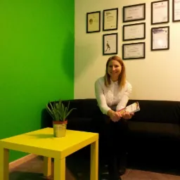 Uśmiechnięta kobieta w białej koszuli siedzi na czarnej sofie w gabinecie z zieloną ścianą, na ścianie wiszą certyfikaty, a obok stoi żółty stolik z rośliną w metalowej doniczce.