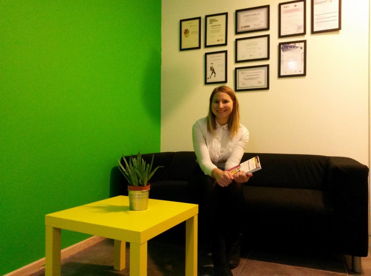 Uśmiechnięta kobieta w białej koszuli siedzi na czarnej sofie w gabinecie z zieloną ścianą, na ścianie wiszą certyfikaty, a obok stoi żółty stolik z rośliną w metalowej doniczce.