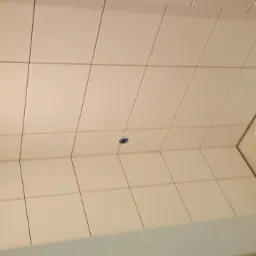 Białe płytki ceramiczne ułożone na ścianie i podłodze łazienki, widoczne niedokończone prace wykończeniowe w rogu pomieszczenia z wystającymi rurami.