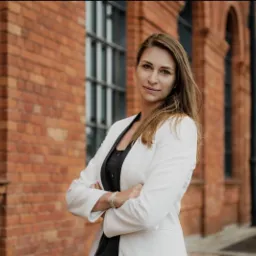Kobieta w białej marynarce, z założonymi rękoma, stoi na tle ceglanego budynku z rzędami okien.