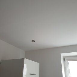 Sławomir Michalski - Biały, gładki sufit z wbudowanym punktowym oświetleniem, fragment szafki kuchennej i okno z roletą w tle. Minimalistyczne wnętrze po remoncie.